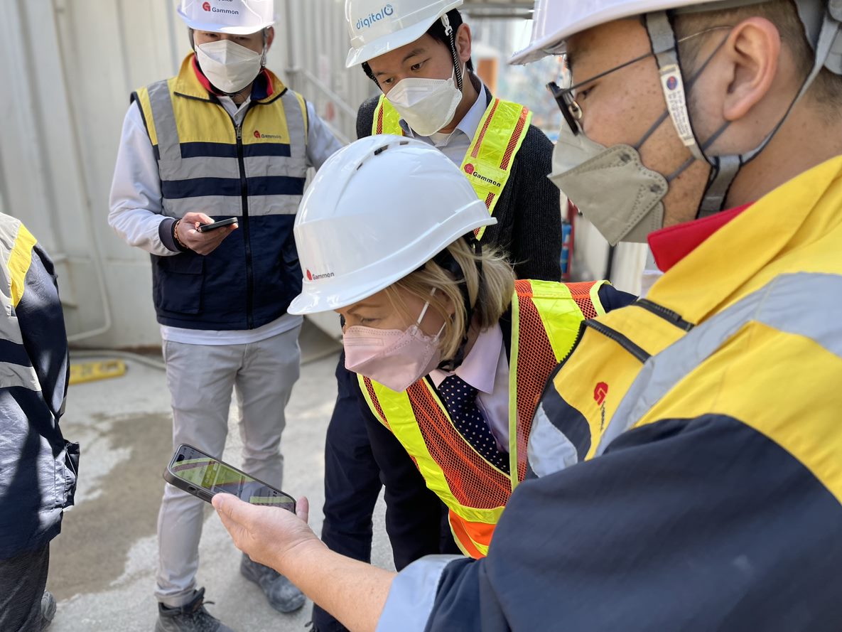 Image of Guildhawk CEO Jurga Zilinskiene MBE on a constructions site in Hong Kong standing with three civil engineers all wearing safety helmets and white hard hats. Jurga is looking at a handheld device an engineer is showing her.  